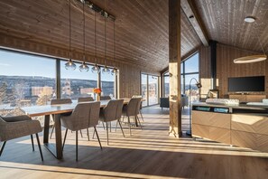A modern interior with a wooden ceiling, large windows, a dining area with chairs, a kitchen with a dishwasher, and a TV.