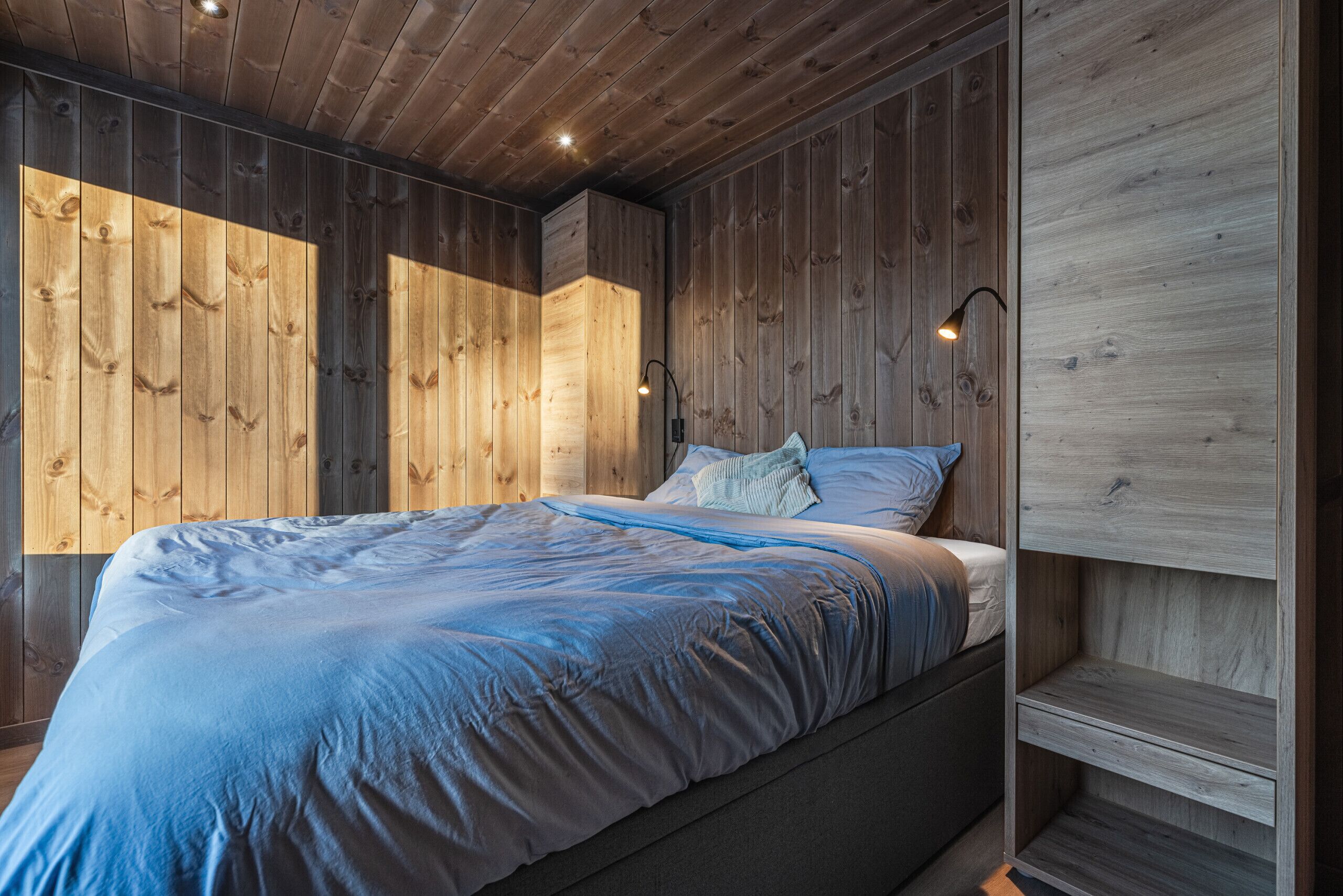 Une chambre à coucher avec un plafond en bois, un lit recouvert d’une couverture bleue et une table de chevet avec une lampe.