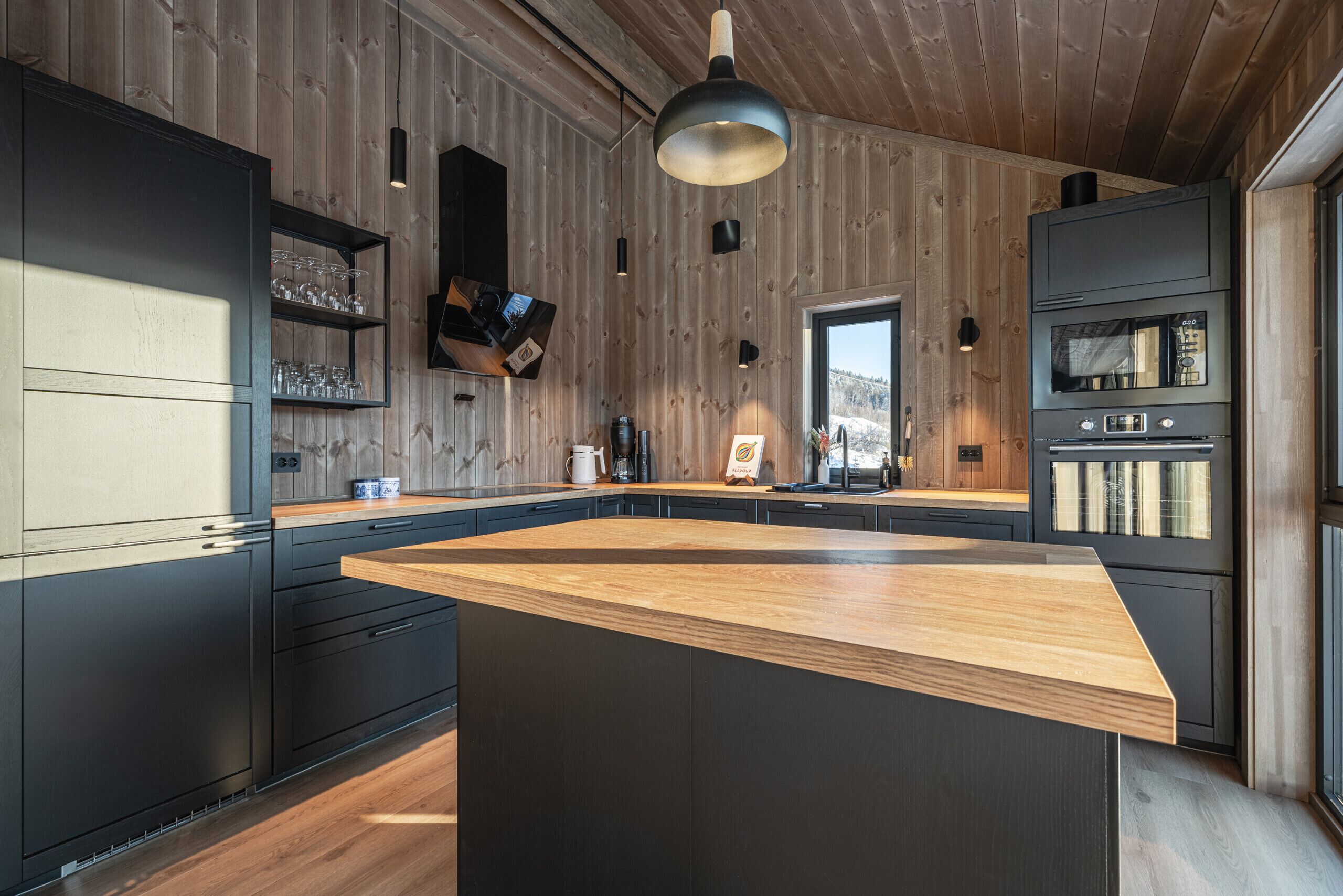 A modern kitchen with a large island, dark cabinetry, wooden walls, and a view of the outdoors.