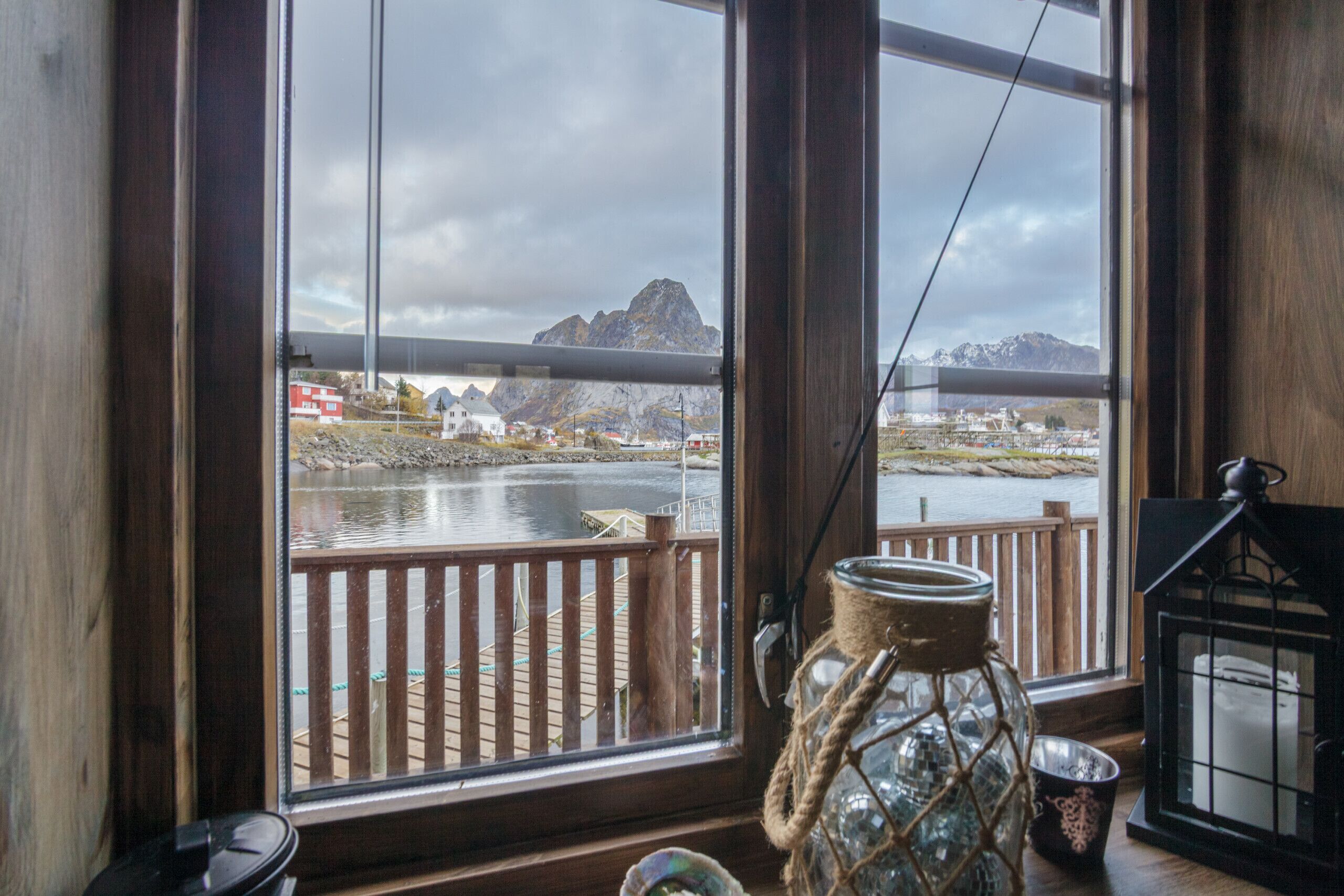 A view through a window with a wooden deck, a fishing rod, a lantern, and a decorative jar.