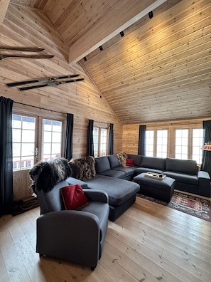 L’intérieur d’une cabane en rondins, avec un canapé d’angle, un petit pouf et un plaid en fourrure.