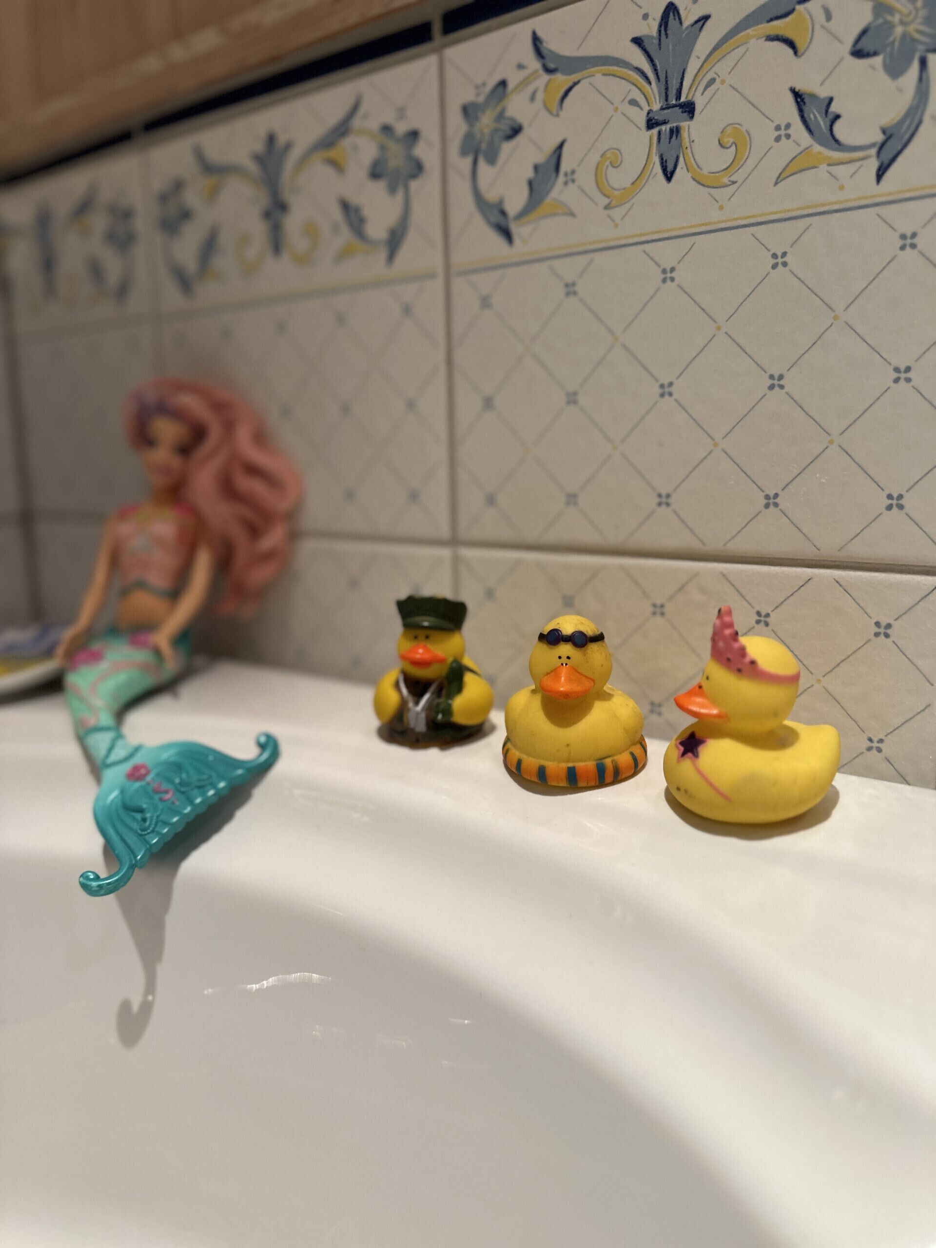 Una bañera con azulejos decorativos, una figurita de sirena y tres patitos de goma.