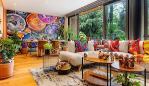 A modern living room with a colorful mural, a sofa with vibrant pillows, a dining area, and a view of greenery outside. - Mexican Folk Art Condesa 3BR, Parking & AC (Mexico City)