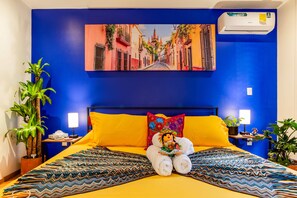 Quarto de hotel com parede azul vibrante, uma cama grande com roupa de cama amarela, uma almofada decorativa e um ar condicionado fixado na parede.