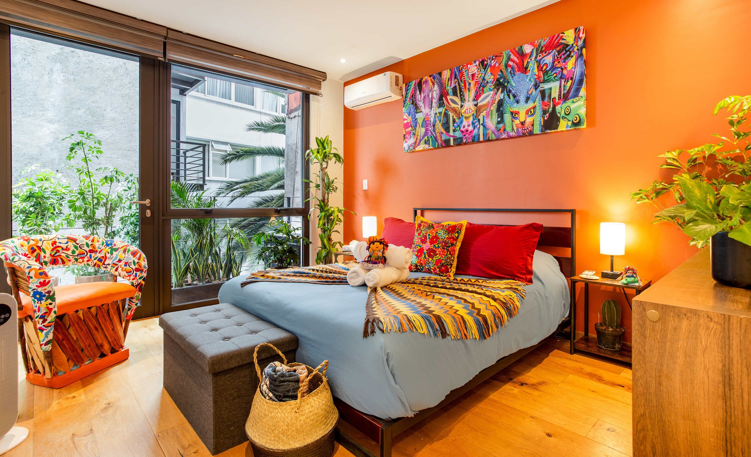 A modern bedroom with a large bed, a colorful wall art piece, a window with a view of greenery, and a cozy seating area with a patterned chair.