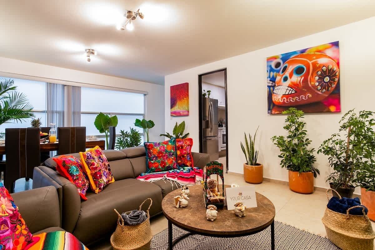 Un salon moderne avec un canapé, une table basse et des coussins décoratifs. On y trouve également des plantes en pot et un tableau accroché au mur.