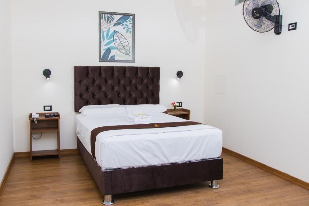 A hotel room with a tufted headboard, a bed with white bedding, a bedside table with a lamp, a wall-mounted fan, and a framed artwork above the bed.