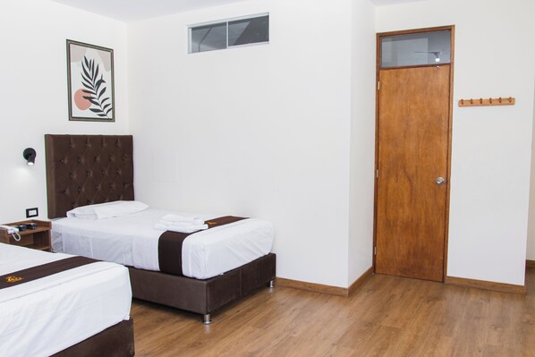 Habitación de hotel con dos camas, una puerta de madera, un ventanal pequeño y un aplique en la pared.
