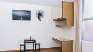 Una habitación con suelo de madera, una pequeña mesa con un banco, un ventilador de pared, un televisor y una ventana con cortinas.