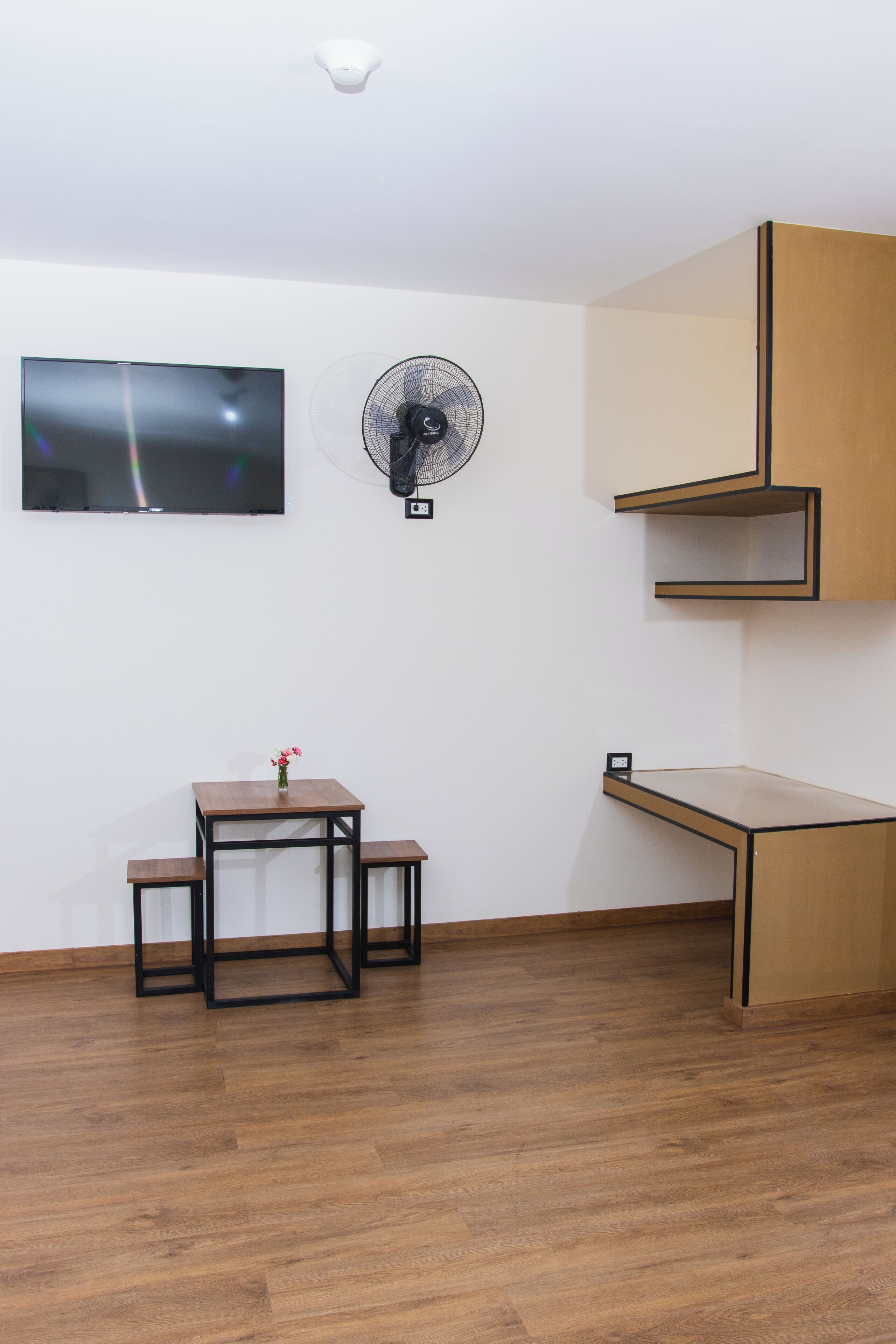 A room with a wooden floor, a TV mounted on the wall, a standing fan, a small table with a vase, and a built-in wooden cabinet.