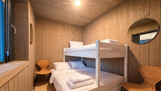 Una cama litera con ropa blanca, una silla de mimbre, un espejo redondo y paredes de madera.