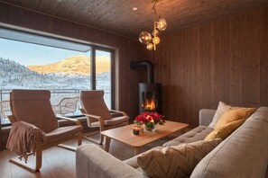 Una habitación acogedora con sofá, sillones, una mesa con flores, chimenea y un ventanal con vista a la montaña.
