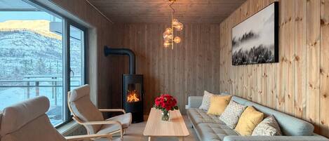 Une pièce chaleureuse avec une cheminée, des murs en bois, un canapé, des fauteuils et une grande fenêtre.