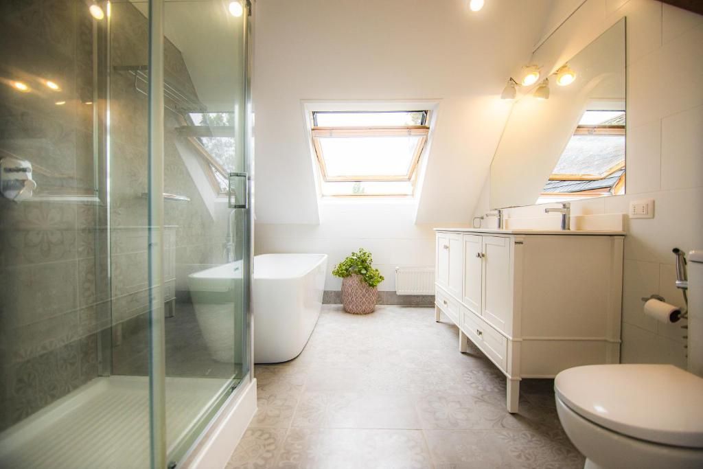 Une salle de bain moderne avec une douche à l’italienne en verre, des toilettes blanches, un lavabo avec un miroir et une fenêtre laissant entrer la lumière naturelle.
