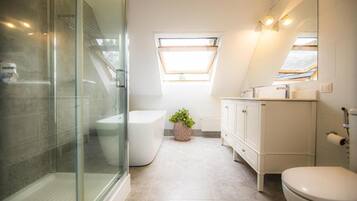 Une salle de bain moderne avec une douche à l’italienne en verre, des toilettes blanches, un lavabo avec un miroir et une fenêtre laissant entrer la lumière naturelle.