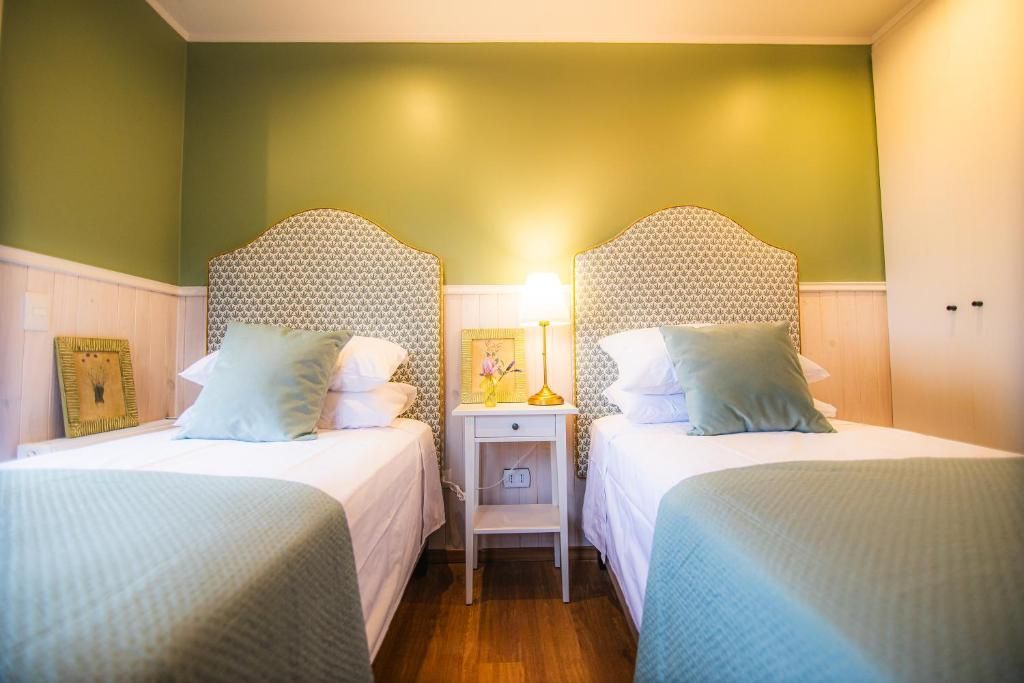 A hotel room with two beds, each with a headboard, a bedside table with a lamp, and a small shelf.