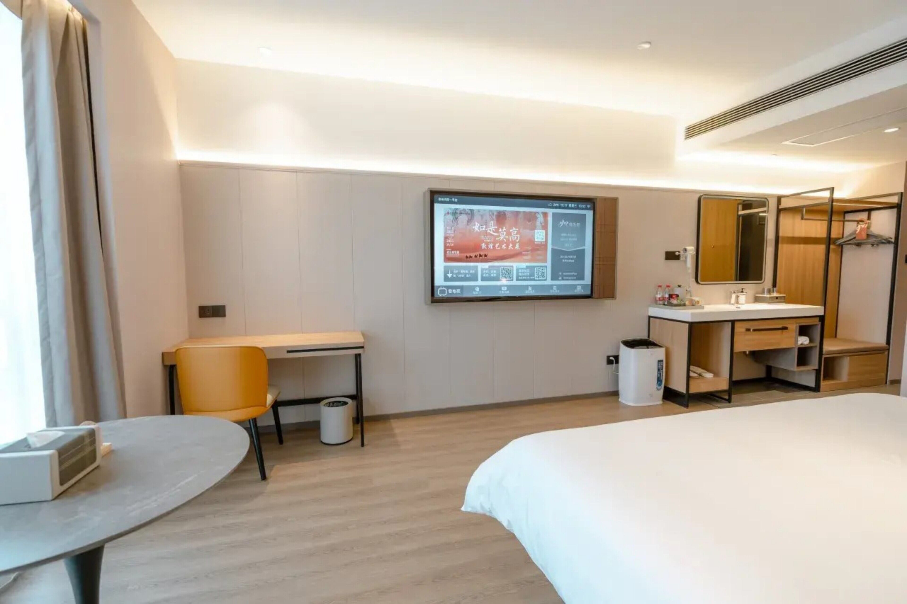 Una habitación de hotel con cama, escritorio, silla, televisor montado en la pared, baño con espejo y cubo de basura.
