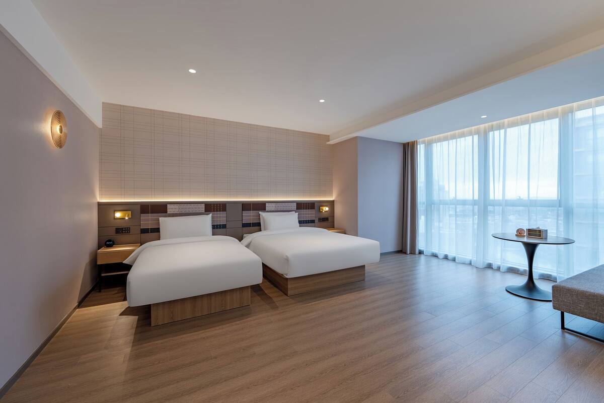 A modern hotel room with two beds, a small table, and a large window.