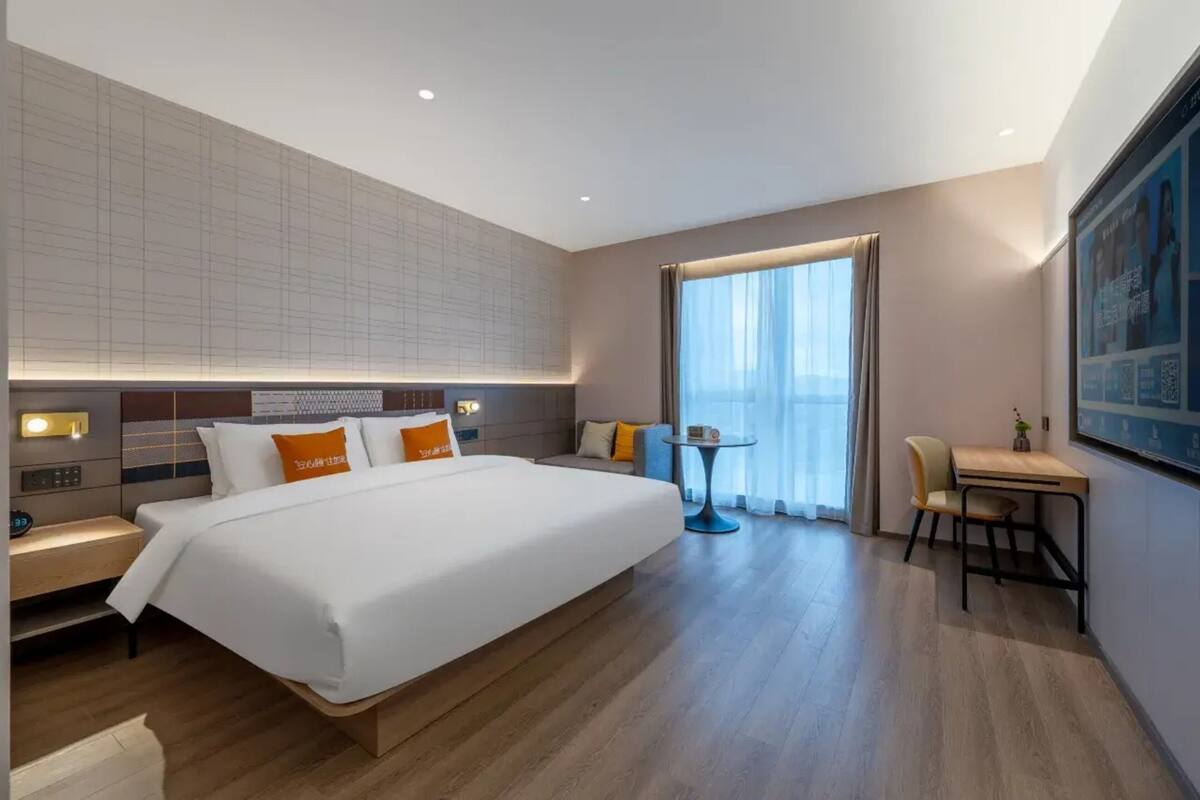 A modern hotel room with a large bed, bedside tables, a small desk, and a view of the city.