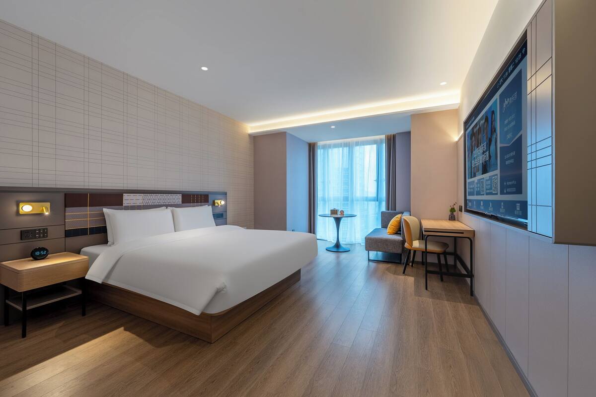 A modern hotel room with a large bed, bedside table, and a desk with a chair.