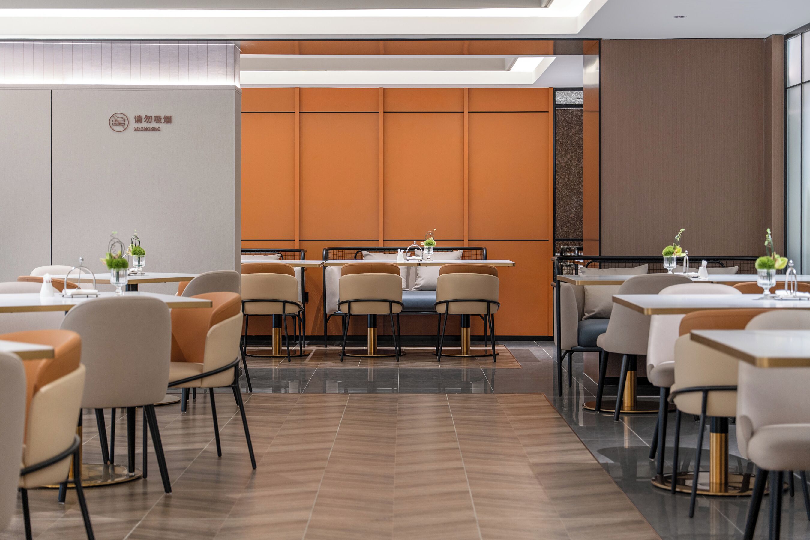 A modern hotel dining area with multiple tables and chairs, a large window, and a wall-mounted sign.