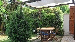 Een houten tafel en stoelen onder een pergola in een tuin met weelderige begroeiing.