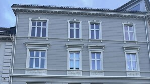 Ein mehrstöckiges Gebäude mit einer symmetrischen Fassade, gleichmäßig angeordneten Fenstern und einem zentralen Eingang mit einer Tür.