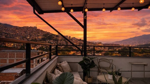 A balcony with a view of a cityscape at sunset, a sofa, a chair, and hanging lights. - Private terrace + panoramic view | Coworking (Medellín)
