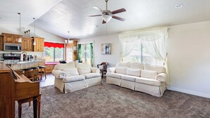 A spacious living area with a piano, a sectional sofa, a ceiling fan, and a kitchen area with wooden cabinets and a microwave.