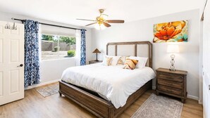 Ein Schlafzimmer mit Bett, Nachttisch, Deckenventilator, Fenster mit Vorhängen und einem Bild an der Wand.