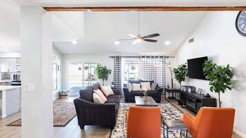 A living room with a sofa, armchairs, a coffee table, and a TV. There is a ceiling fan, a clock, and a plant.