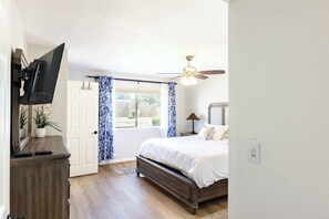 A bedroom with a bed, a flat-screen TV mounted on a dresser, a ceiling fan, and a window with curtains. - Sunset Springs - 4BR Home w/ Private Pool, Hot Tub (Phoenix)