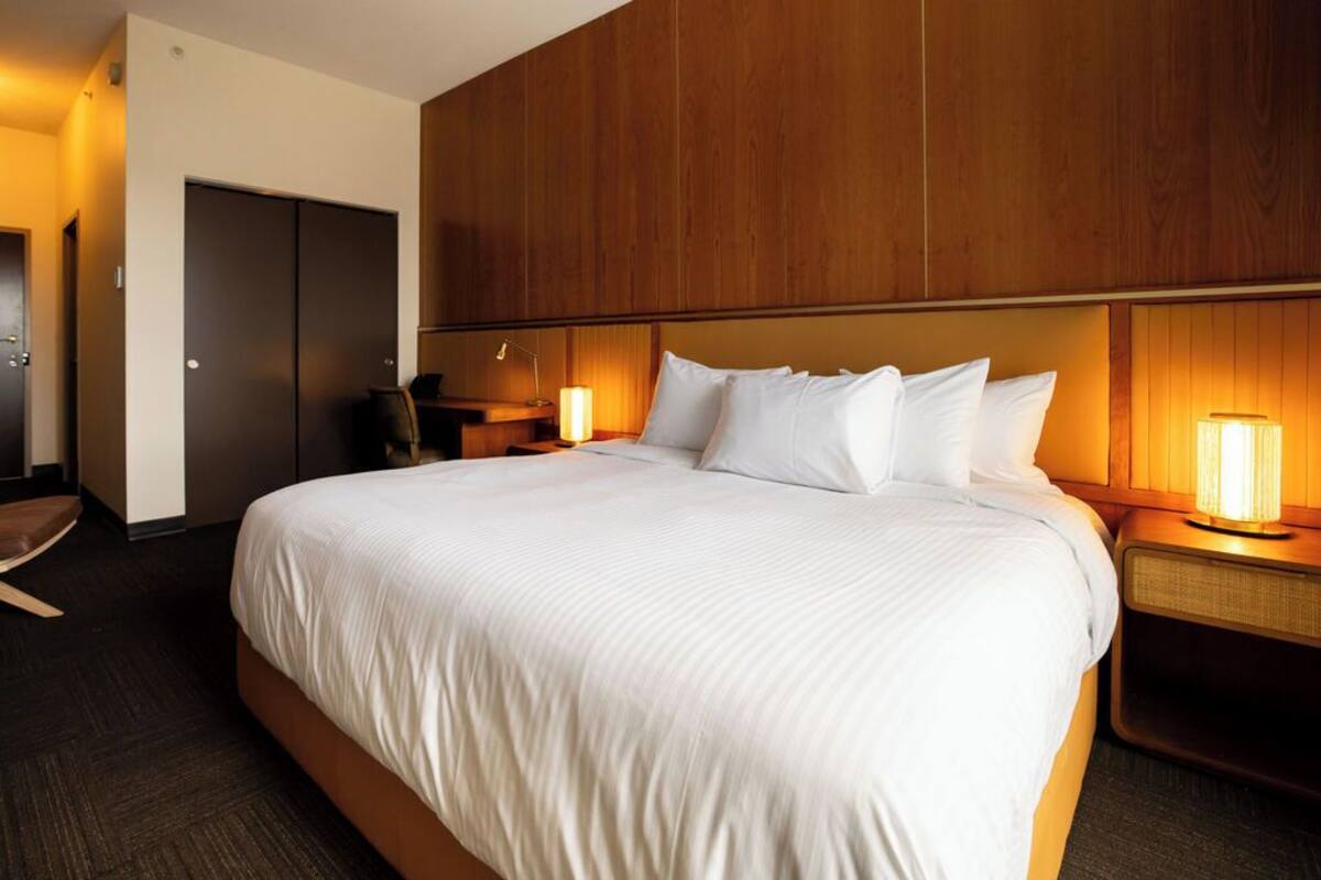 Habitación de hotel con una cama grande, cabecero de madera, dos lámparas de noche, un armario y un escritorio con silla.