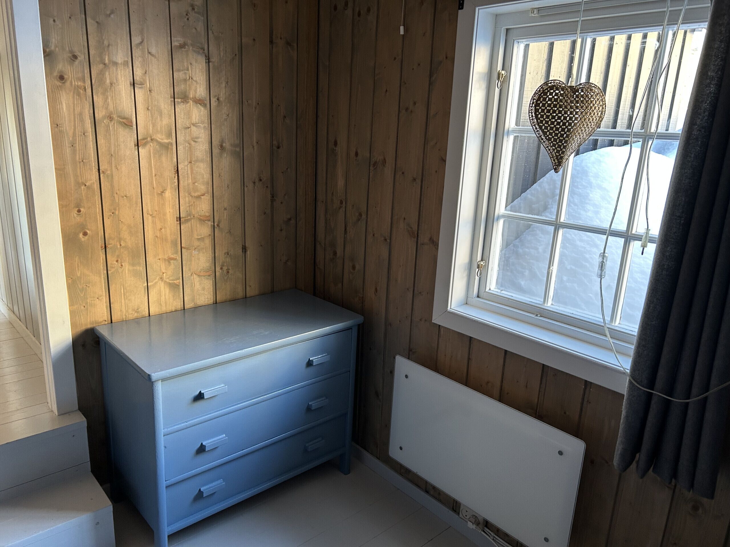 A room with wooden walls, a window with a heart-shaped decoration, a radiator, and a blue dresser.
