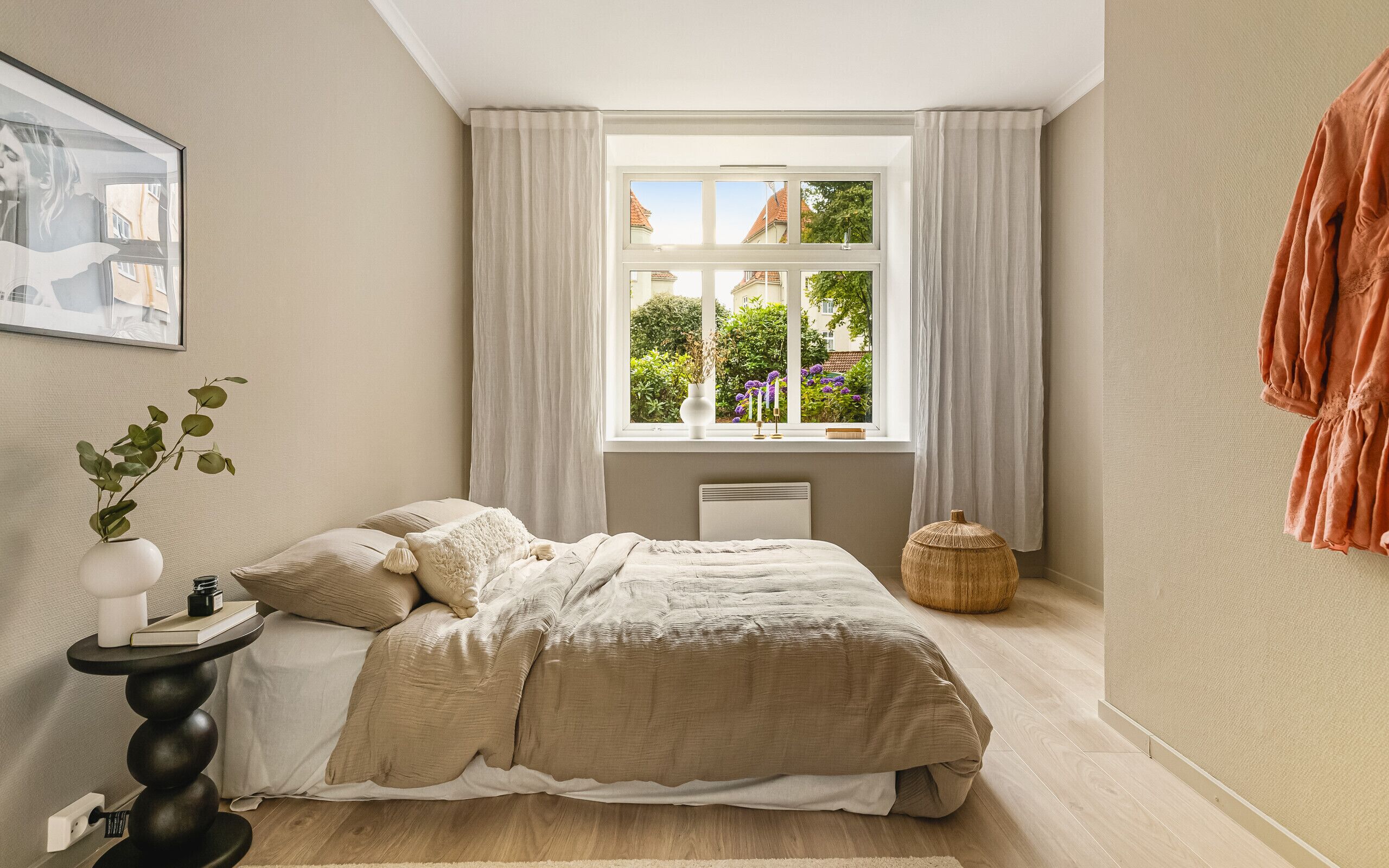 A bedroom with a bed, a bedside table with a plant, a window with curtains, and a radiator.