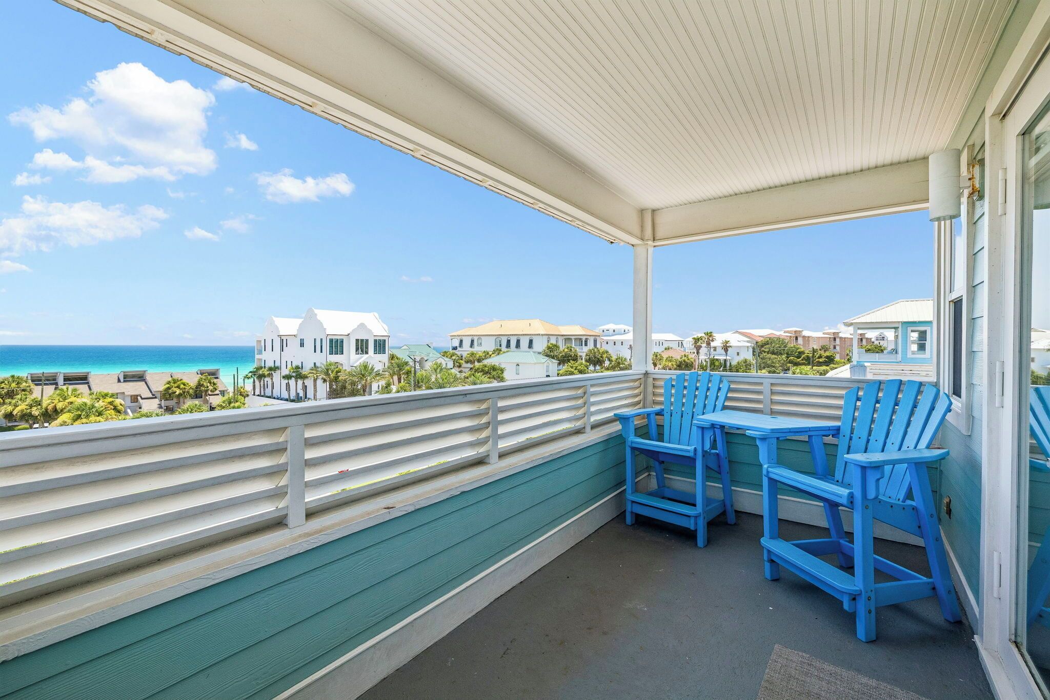 A covered balcony with blue chairs, a view of the ocean, and buildings in the background.