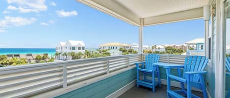 A covered balcony with blue chairs, a view of the ocean, and buildings in the background.