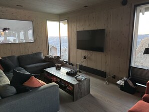 Un salon doté d’un grand téléviseur à écran plat fixé au mur, d’une table basse en bois, d’un canapé et d’une fenêtre donnant sur un paysage enneigé.