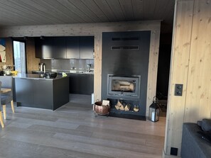 A modern kitchen with a central island, a built-in wood stove, and a dining area with a wooden ceiling.