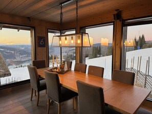 A dining area with a wooden table and chairs, large windows with a snowy mountain view, and a hanging light fixture.