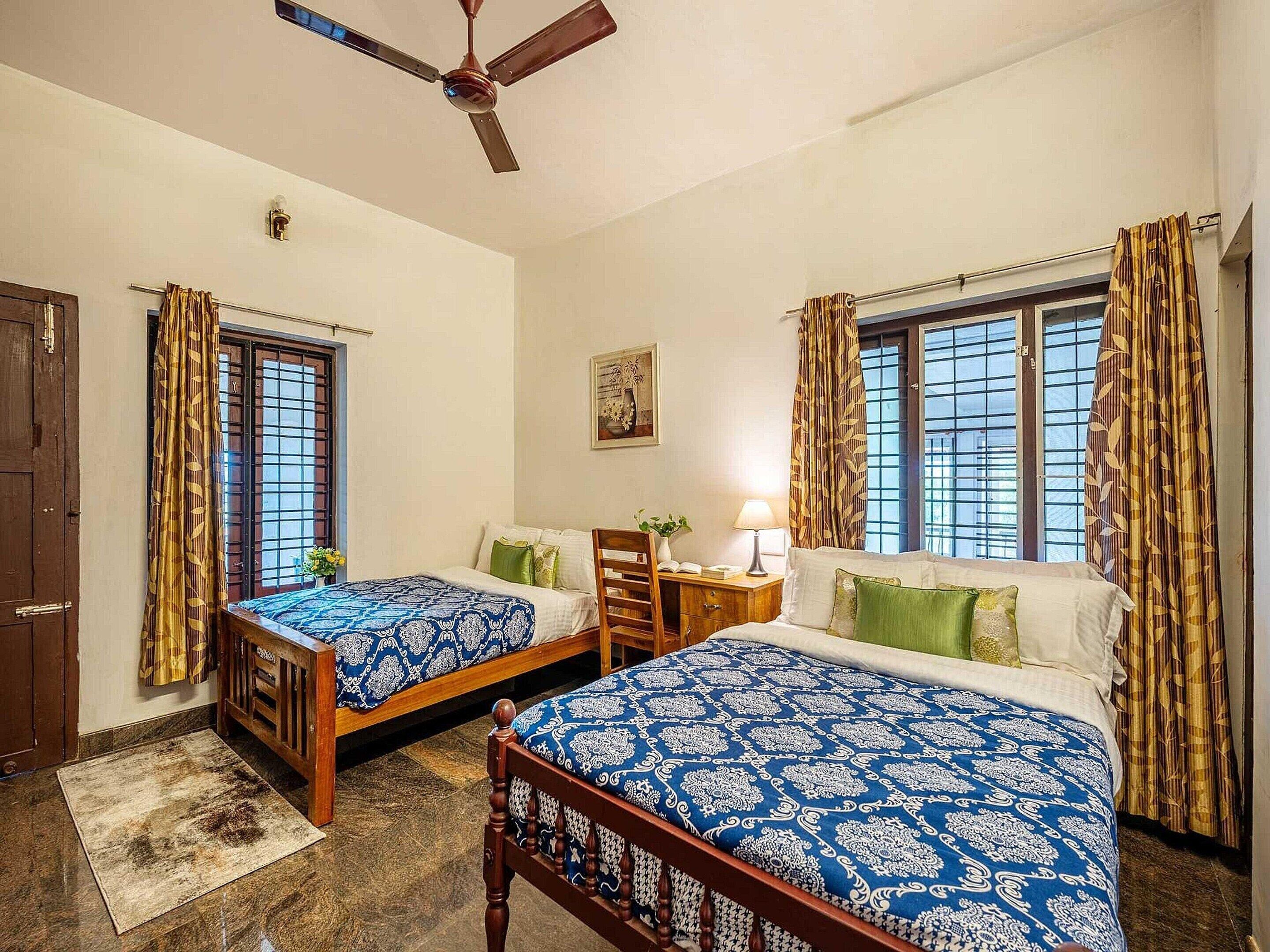 A room with two beds, a desk, a chair, a ceiling fan, and a window with curtains.