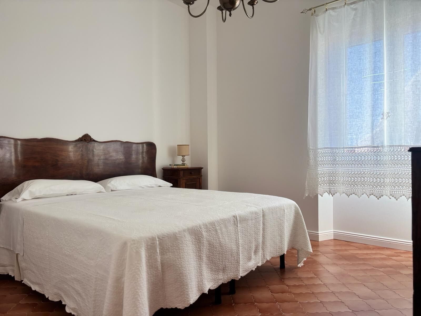 Un dormitorio con una cama de madera, una mesita de noche, una lámpara y una ventana con cortina de encaje.