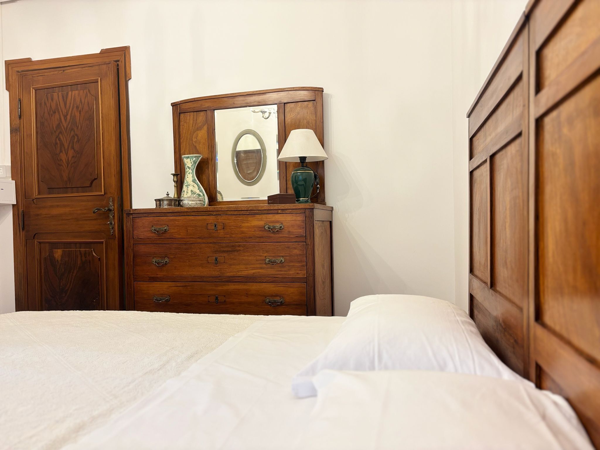A bedroom with a wooden dresser, a mirror, a lamp, and a bed with white bedding.