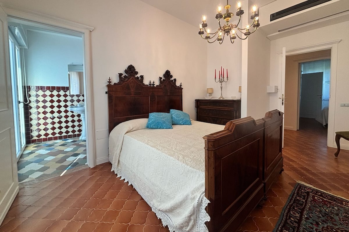 A bedroom with a wooden bed, a chandelier, a patterned floor, and a view of the hallway.