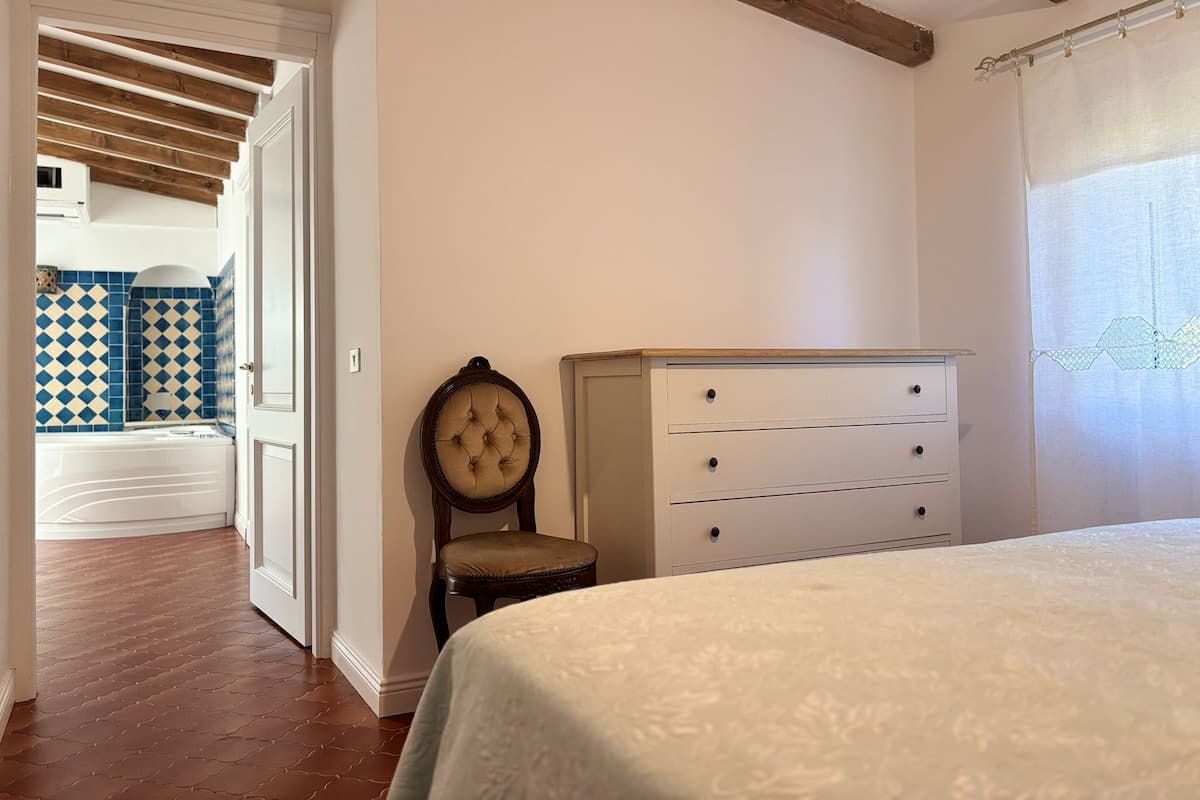 A bedroom with a bed, a dresser, a chair, and a view of a bathroom with patterned tiles.