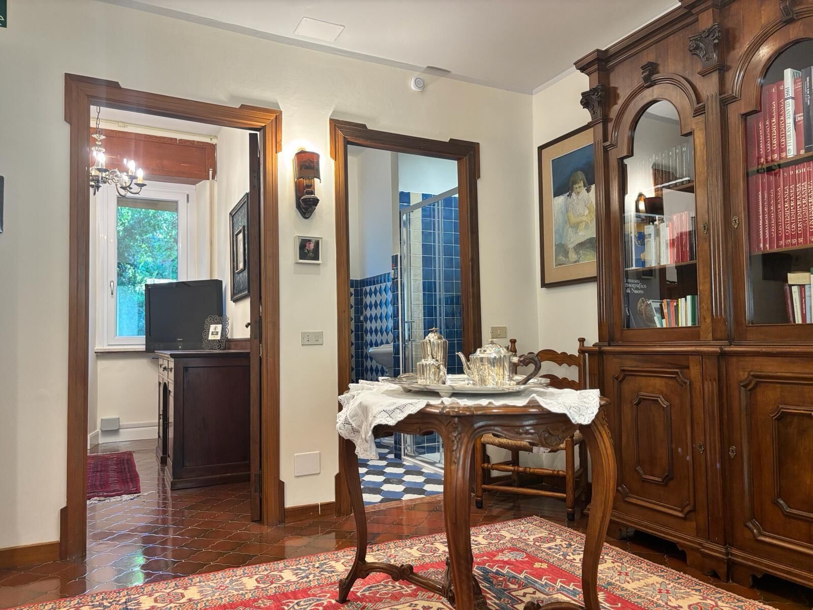 A dining area with a table set for two, a wooden cabinet with books, and a patterned rug.