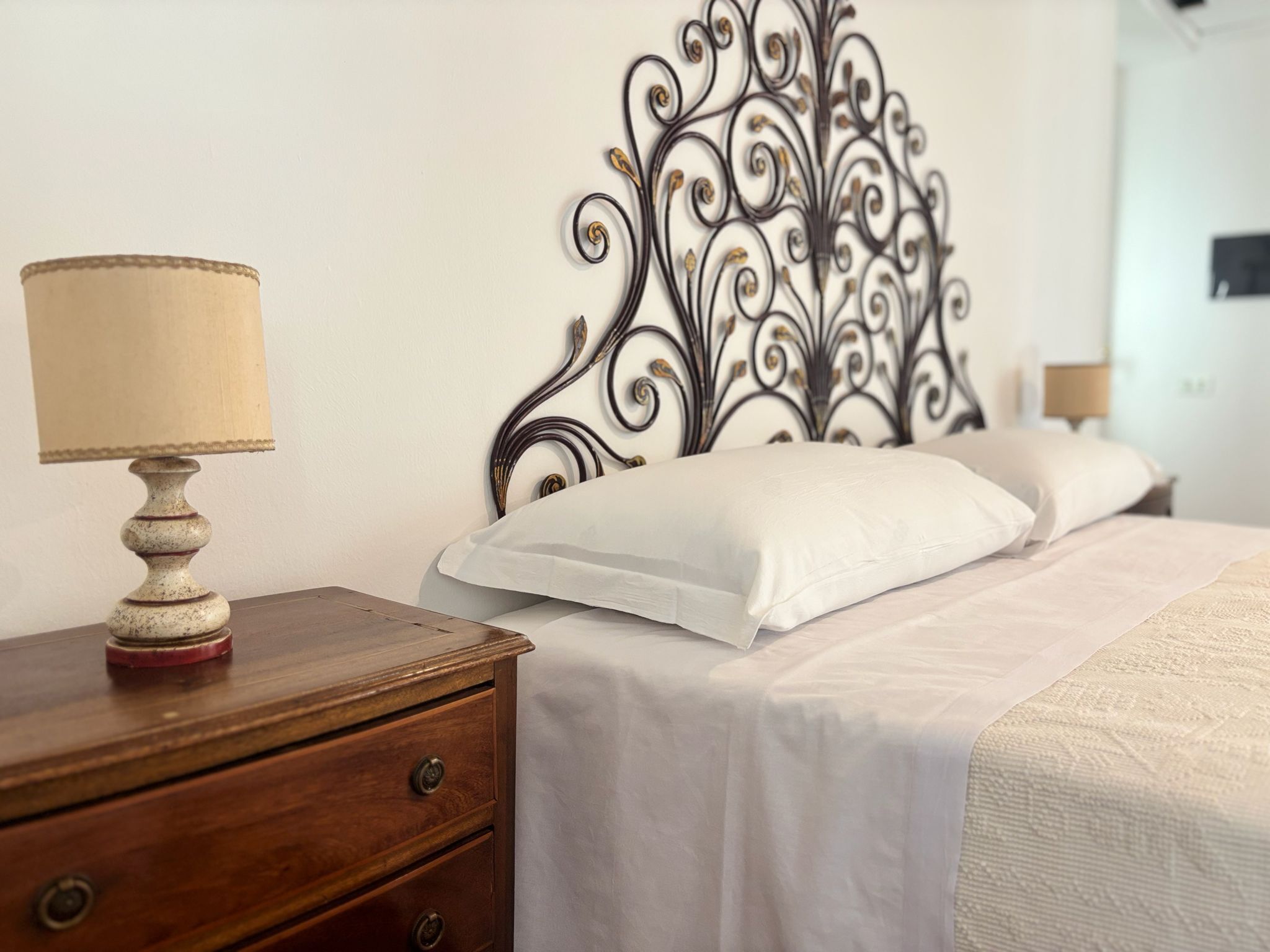 Una cama bien hecha con ropa blanca, una mesita de noche de madera con una lámpara y un cabecero decorativo de metal.