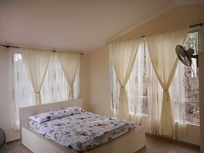A bedroom with a bed, two windows with curtains, and a standing fan. - Girardot Cabañas - Balcón de Guabinal (Girardot)