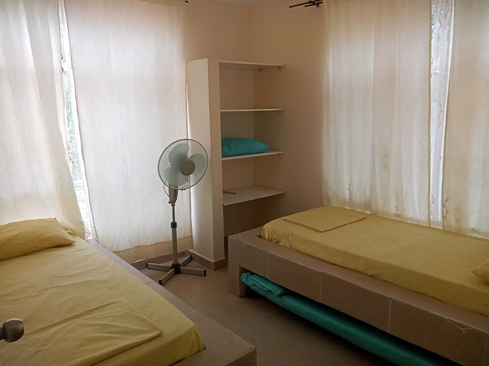 Quarto com duas camas, um ventilador e um guarda-roupa com prateleiras.