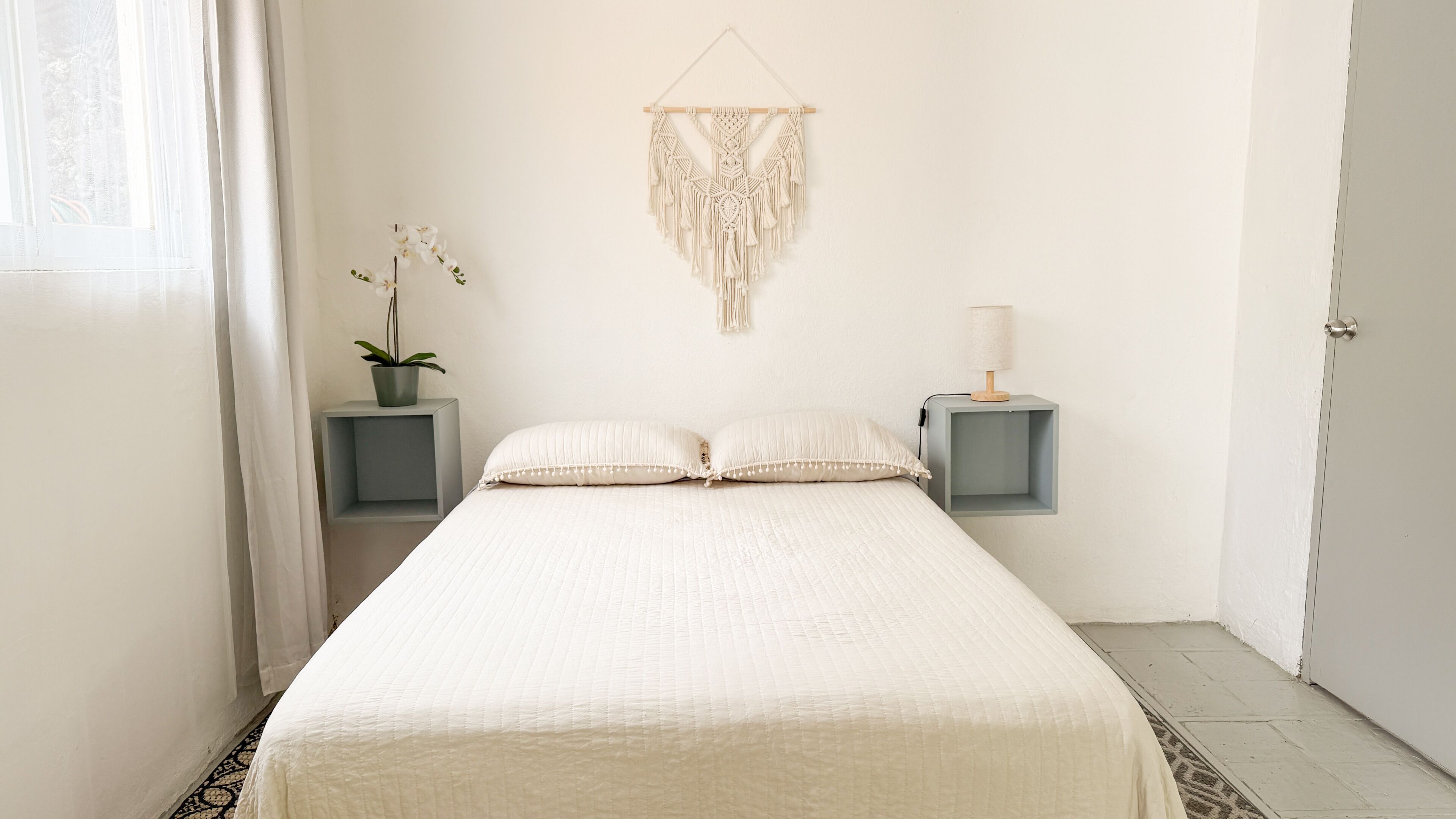 Un dormitorio minimalista con cama, dos mesitas de noche, un colgador de macramé, una planta en maceta y una lámpara.